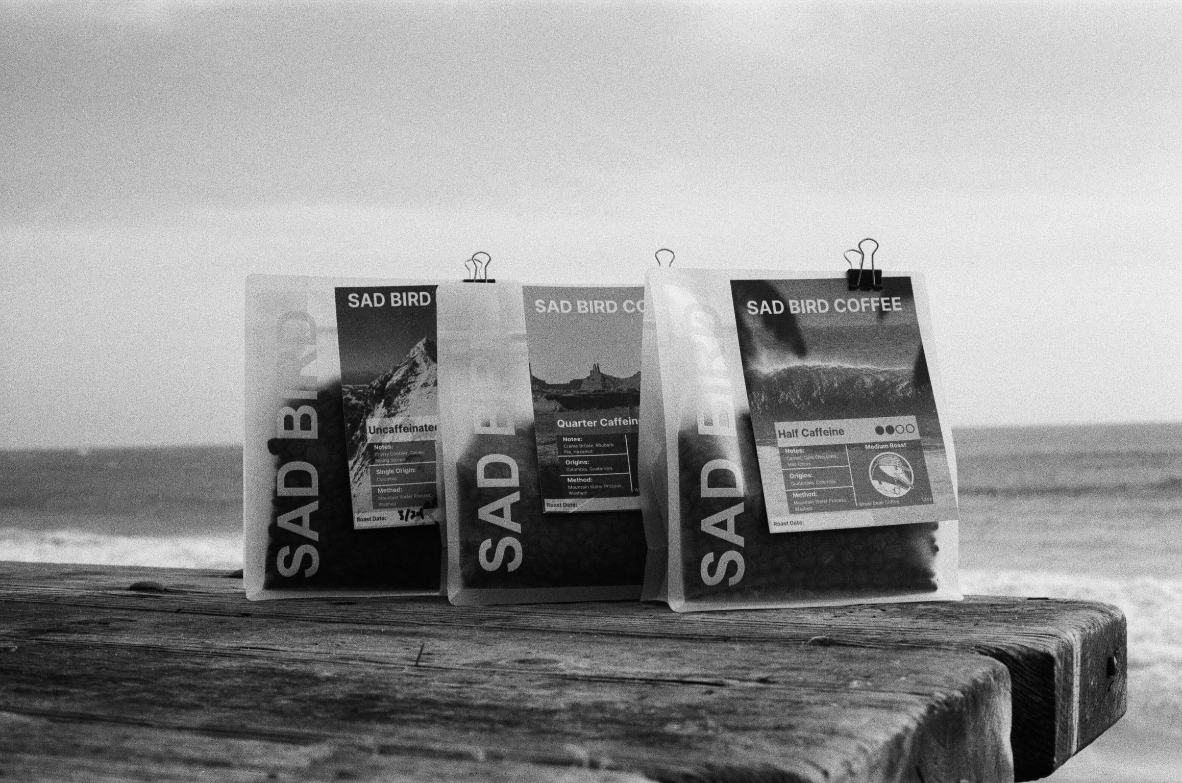 Three packages of Sad Bird Coffee on a wooden surface with a beach background